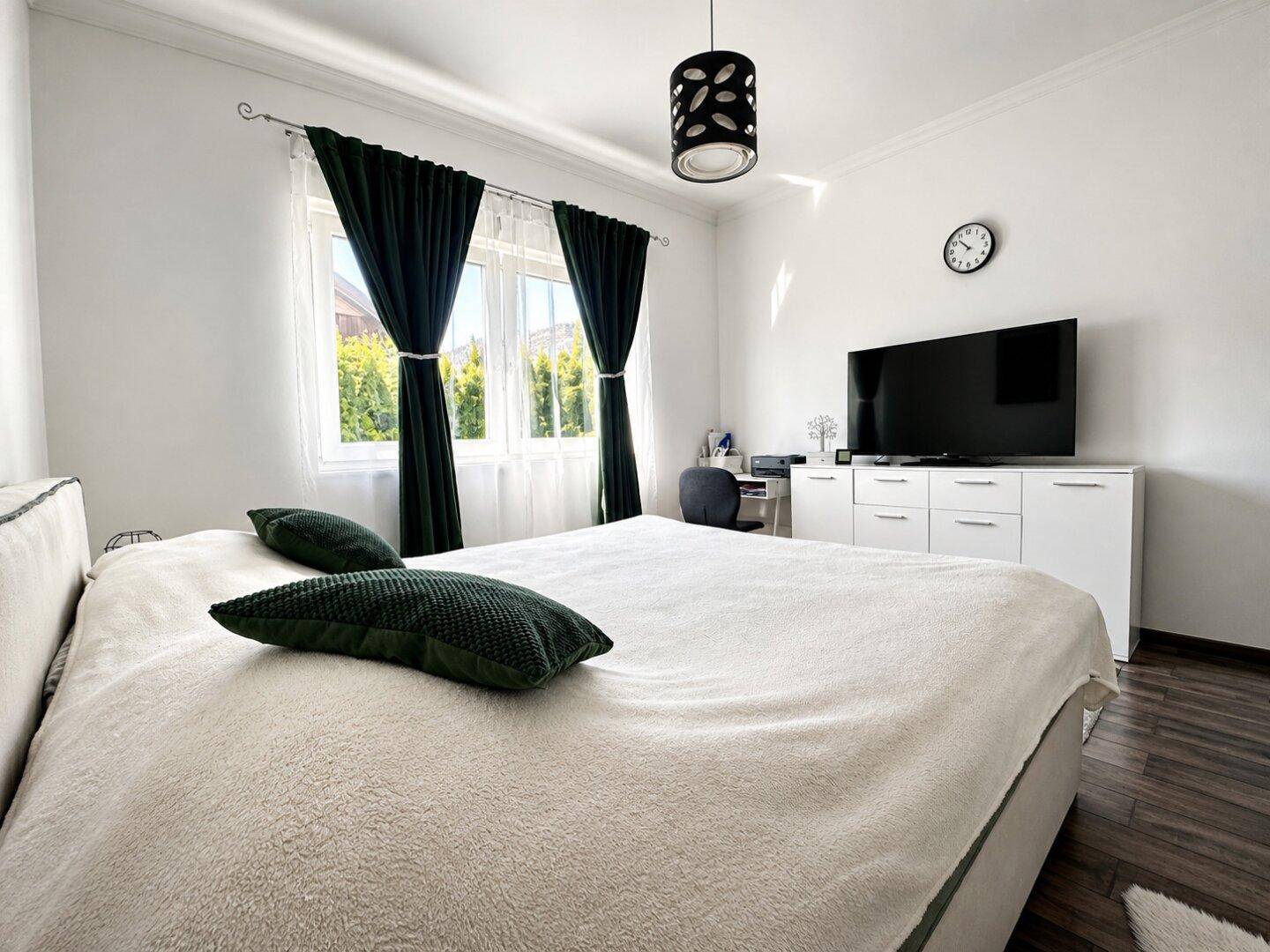 Helles Schlafzimmer mit Doppelbett, Schreibtisch und Fernseher, ergänzt durch dunkle Vorhänge.