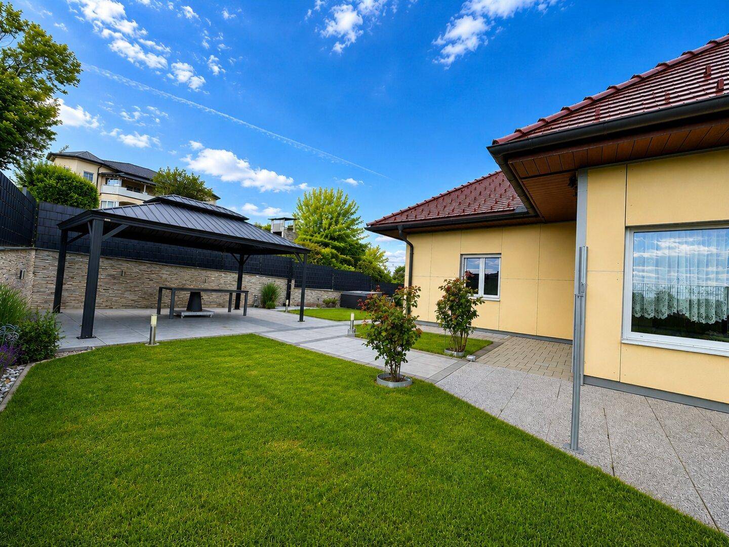 Gepflasterter Gartenbereich mit moderner Pergola und Sitzgelegenheiten, umgeben von Rasen.