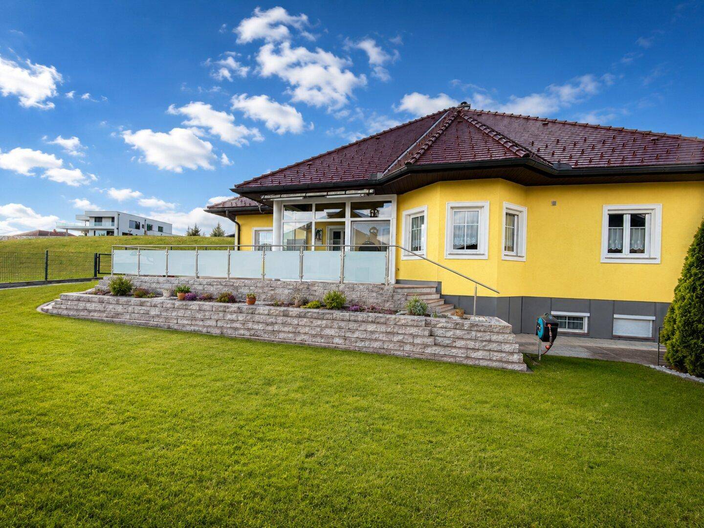 Ansicht des Hauses mit gelber Fassade, gepflegtem Rasen und einer Terrasse mit Glasgeländer.