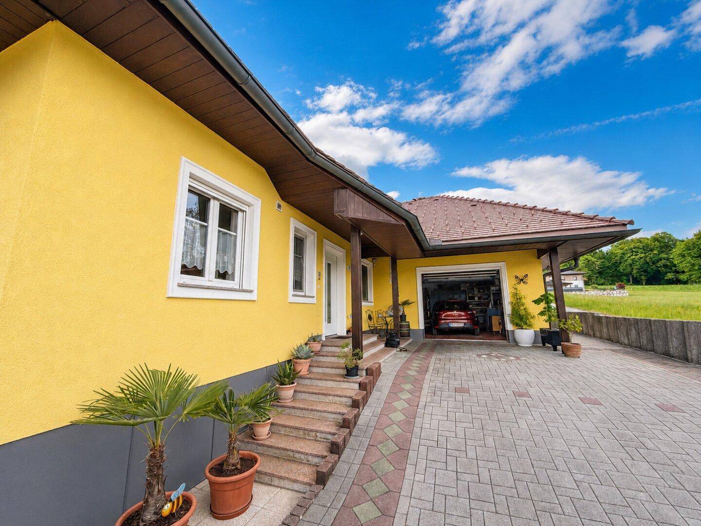 Vorderansicht des Hauses mit gelber Fassade, gepflasterter Auffahrt und integrierter Garage.