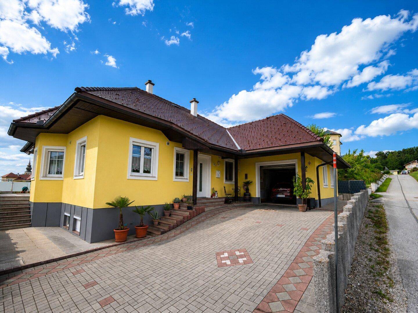 Ansicht des Hauses mit gelber Fassade, gepflasterter Auffahrt und Zugang zur Garage.