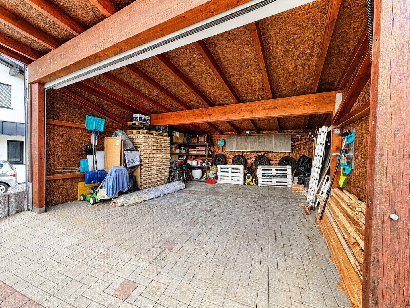 Überdachter Stellplatz oder Carport mit Holzbalken und Stauraum für Gartengeräte.