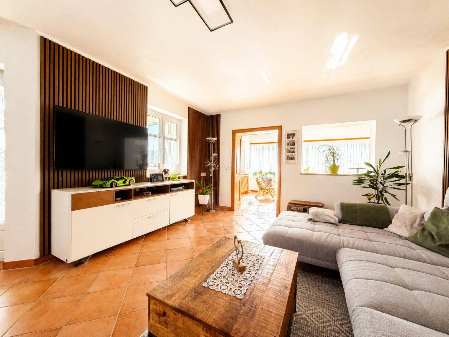 Helles Wohnzimmer mit großem Sofa, Fernseher und Zugang zu einem weiteren hellen Raum.