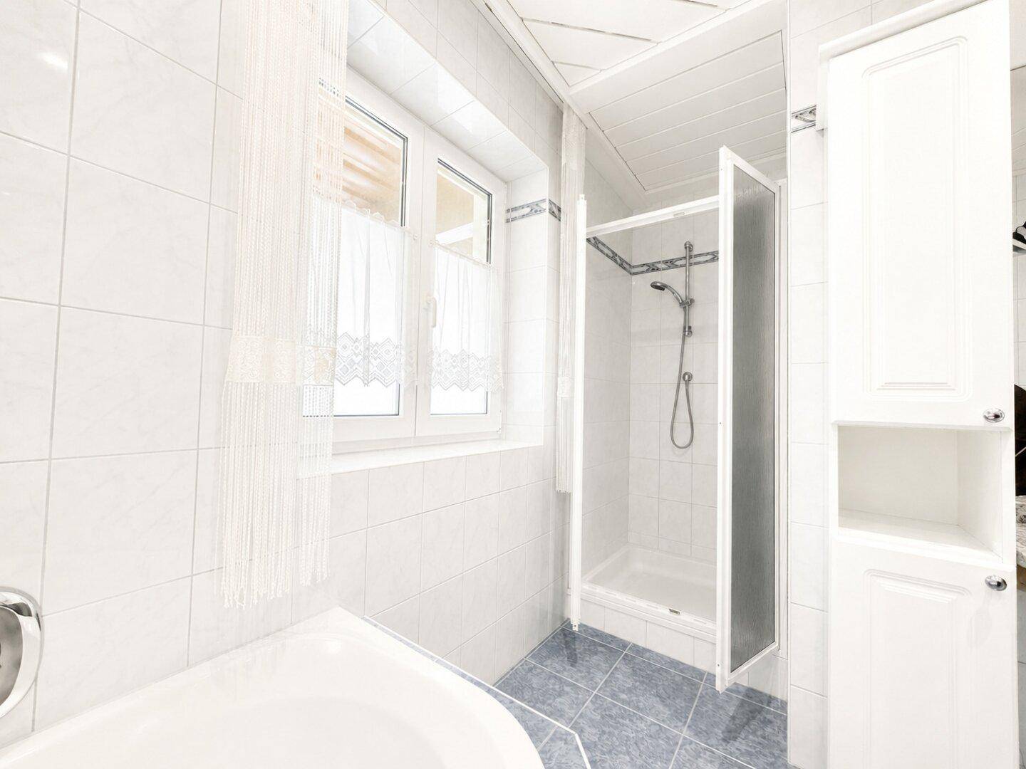 Badezimmer mit Badewanne, Duschkabine, weißen Wandfliesen und Fenster.