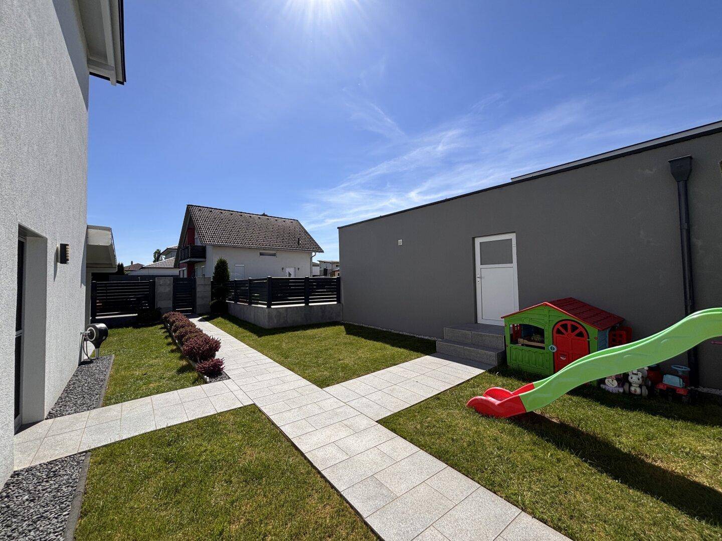 Seitlicher Blick in den Garten mit gepflastertem Weg, Rasenfläche und einem kleinen Kinderspielhaus.