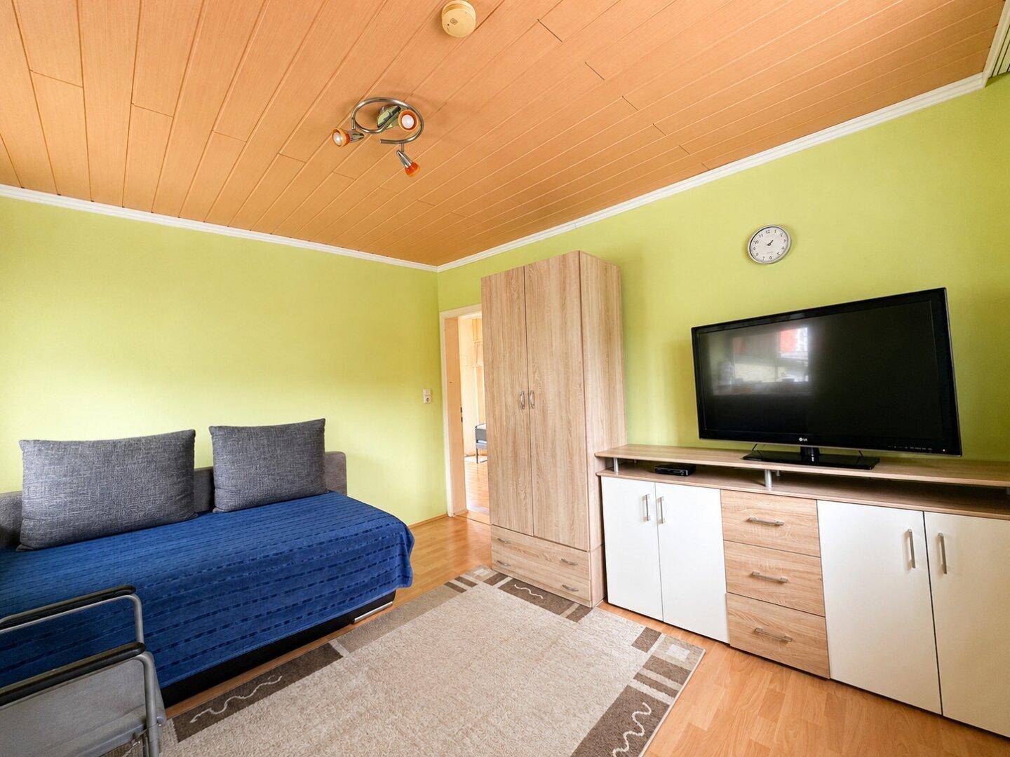 Wohnzimmer mit hellgrünen Wänden, Holzdecke und einem Sofa sowie TV-Möbeln.