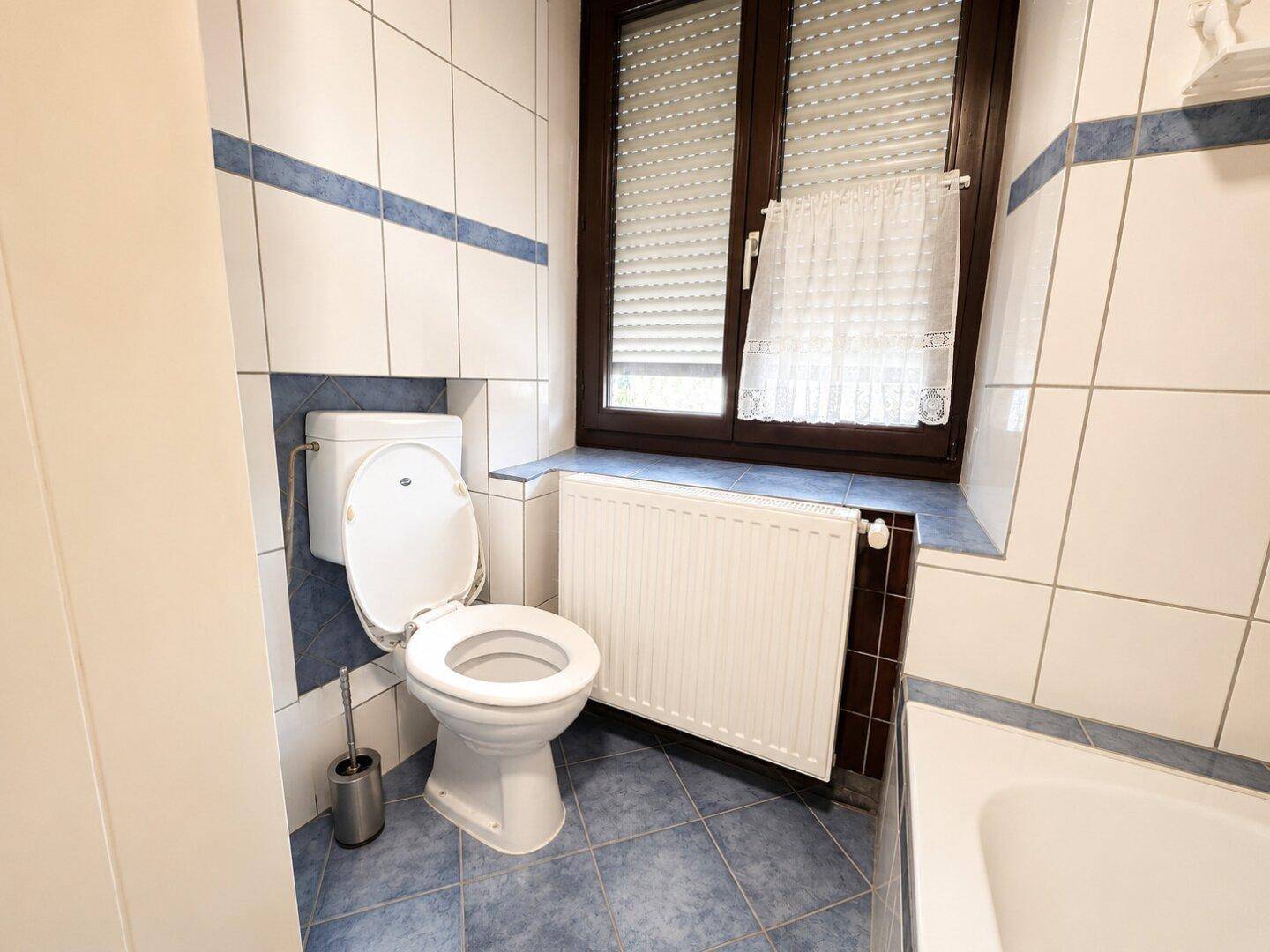 Toilette im Badezimmer mit weißen Fliesen, Heizkörper und Fenster.