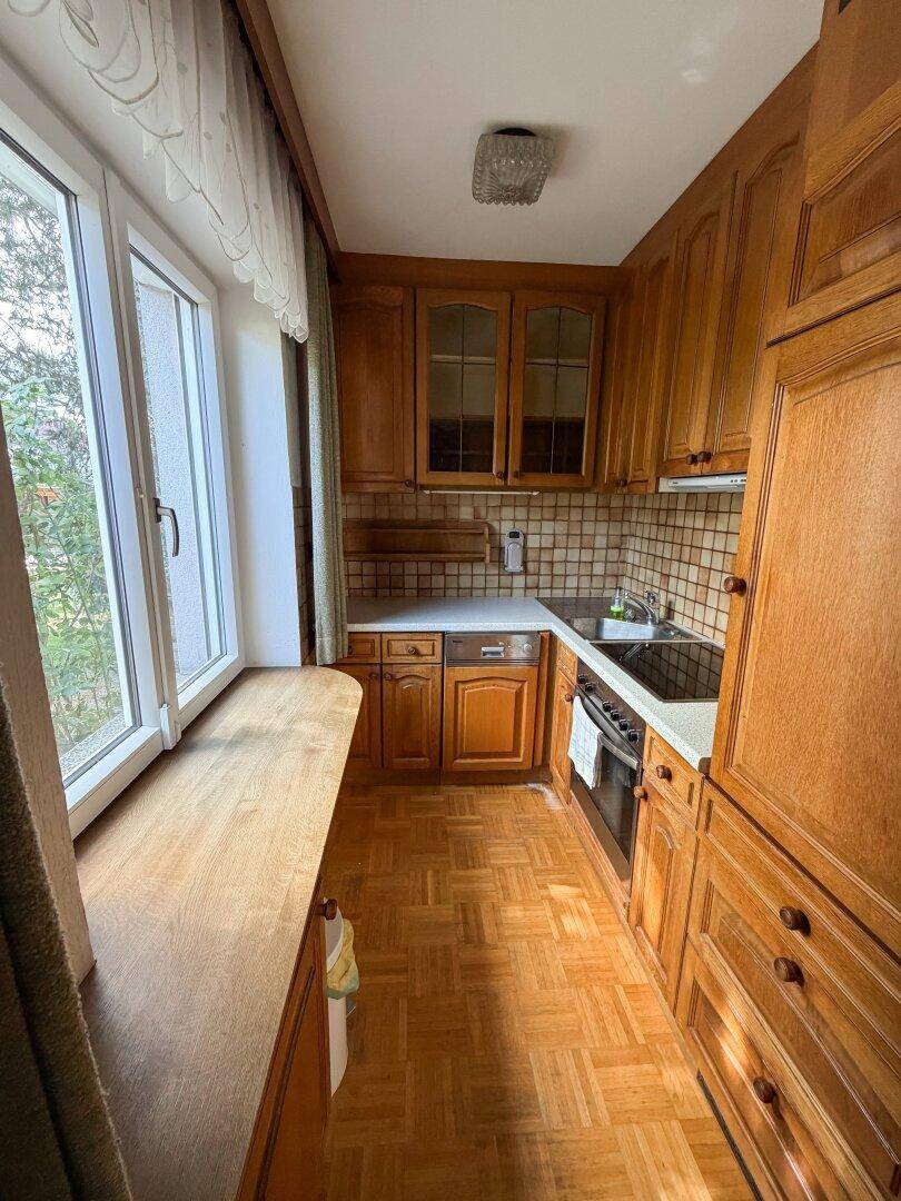 Schmale Küche mit Holzschränken, Parkettboden und einem Fenster, das viel Tageslicht hereinlässt.