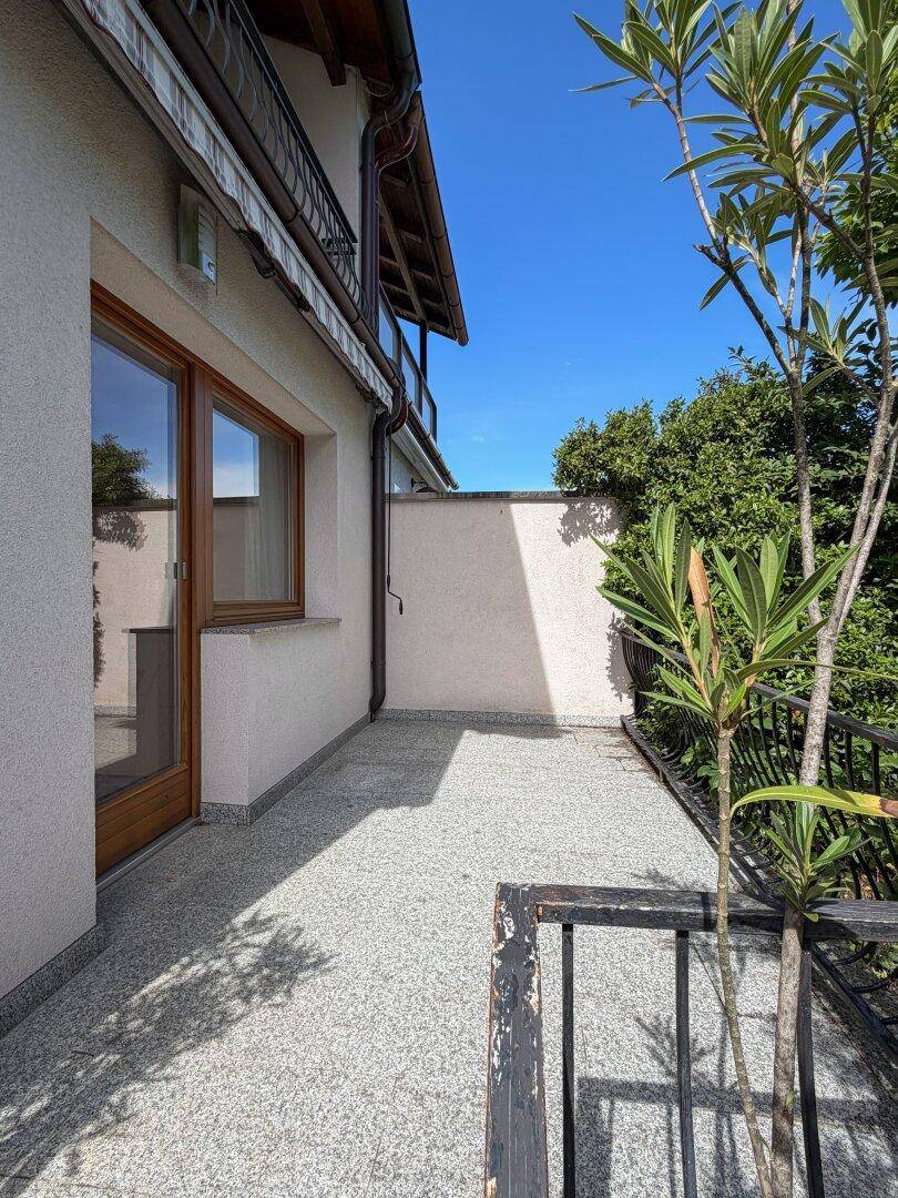 Eine kleine Terrasse mit grauen Fliesen und einer niedrigen Mauer, umgeben von Grünpflanzen.