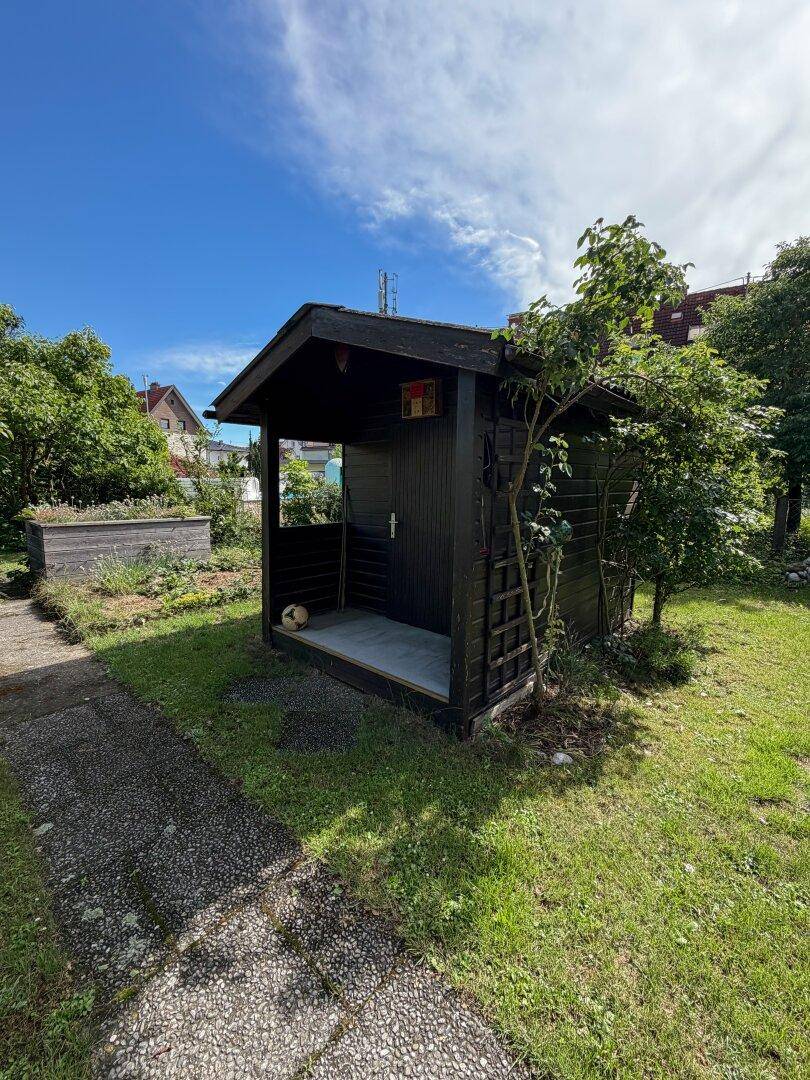 Kleiner Gartenschuppen in einer grünen Umgebung mit einem Hochbeet im Hintergrund.