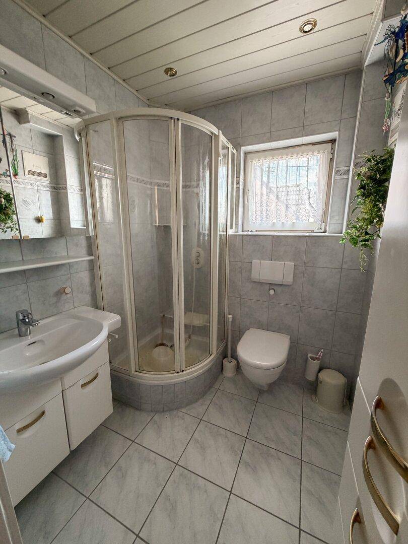 Badezimmer mit Duschkabine, WC, Waschbecken und grauen Fliesen.