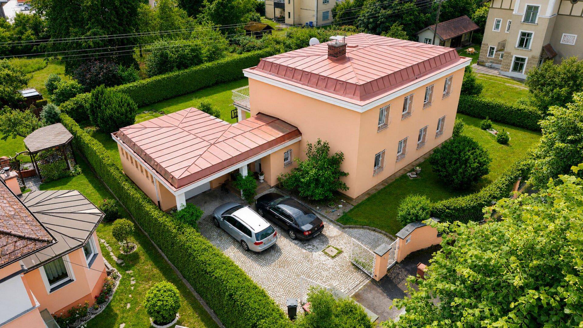 Luftaufnahme des Hauses mit großem Garten, Carport und gepflasterter Einfahrt, umgeben von Grün.