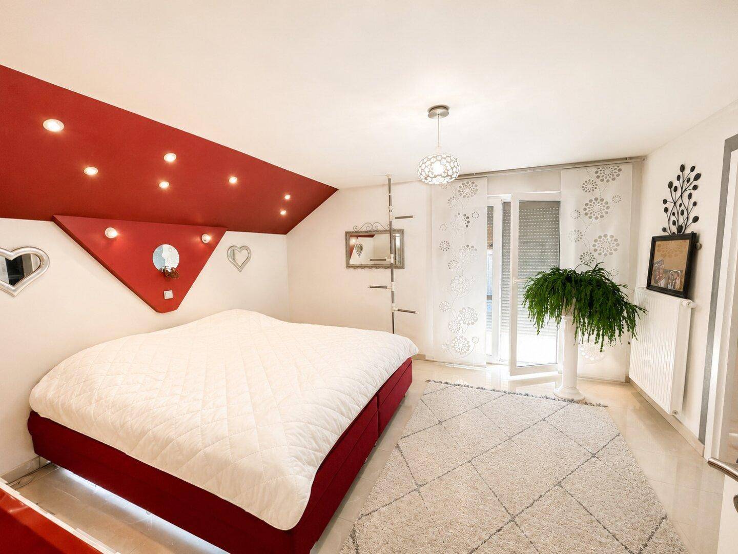 Helles Schlafzimmer im Dachgeschoss mit roter Akzentwand, Doppelbett und Zugang zum Balkon.