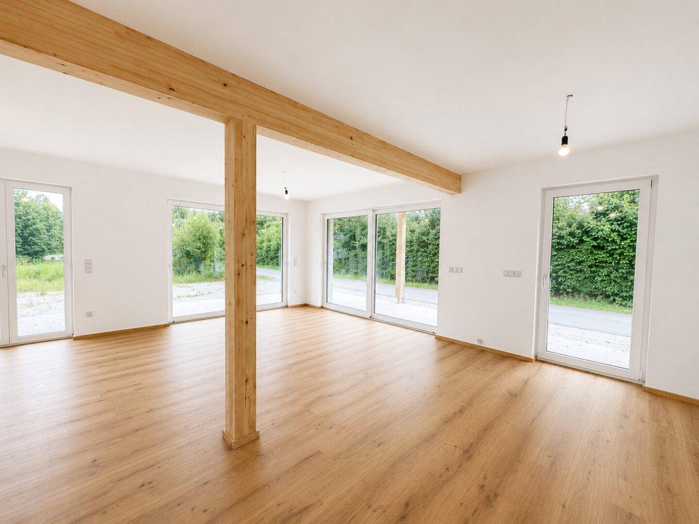 Lichtdurchfluteter Wohnraum mit großen Fenstern, Holzbalken und Blick ins Grüne.
