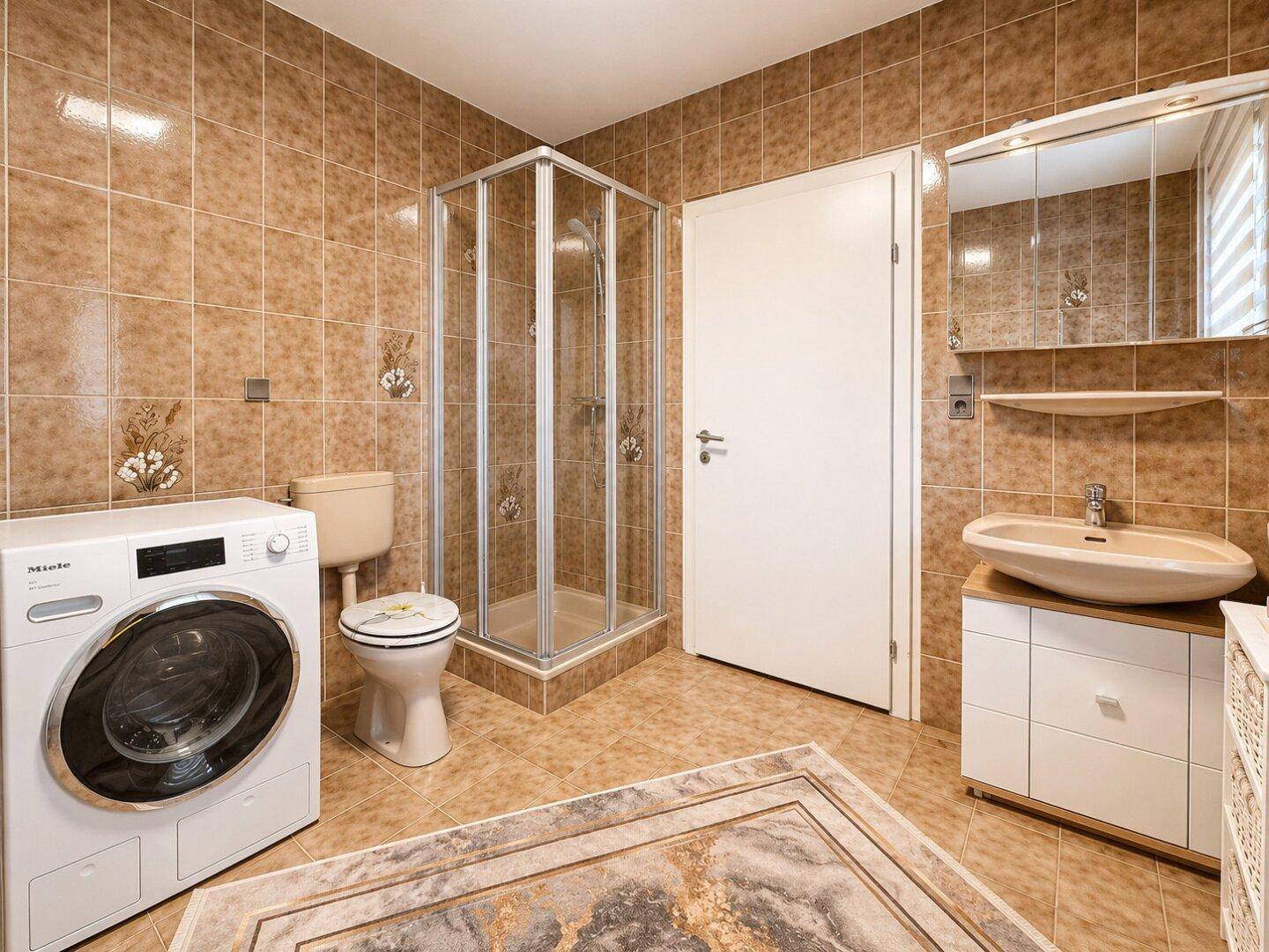 Badezimmer mit braunen Fliesen, Duschkabine, Toilette und einer Waschmaschine.