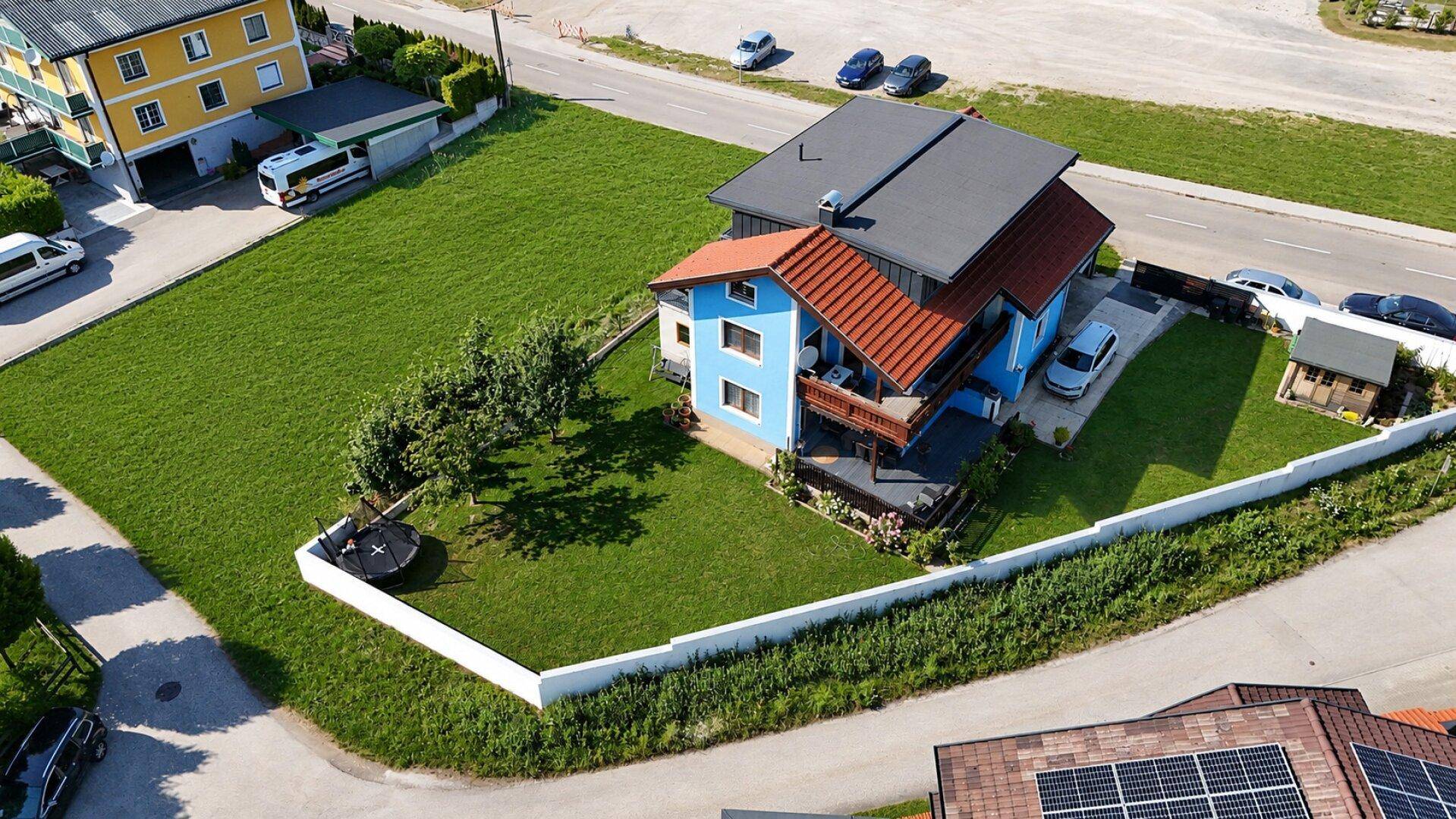 Luftaufnahme eines blauen Hauses mit großem Garten, Trampolin und Nebengebäude in ländlicher Umgebung.