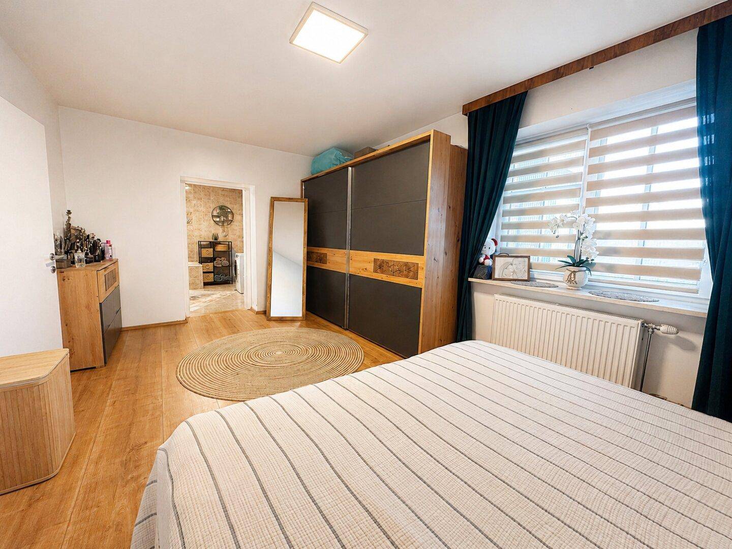 Schlafzimmer mit Fenster, Heizkörper und Zugang zu einem angrenzenden Badezimmer.
