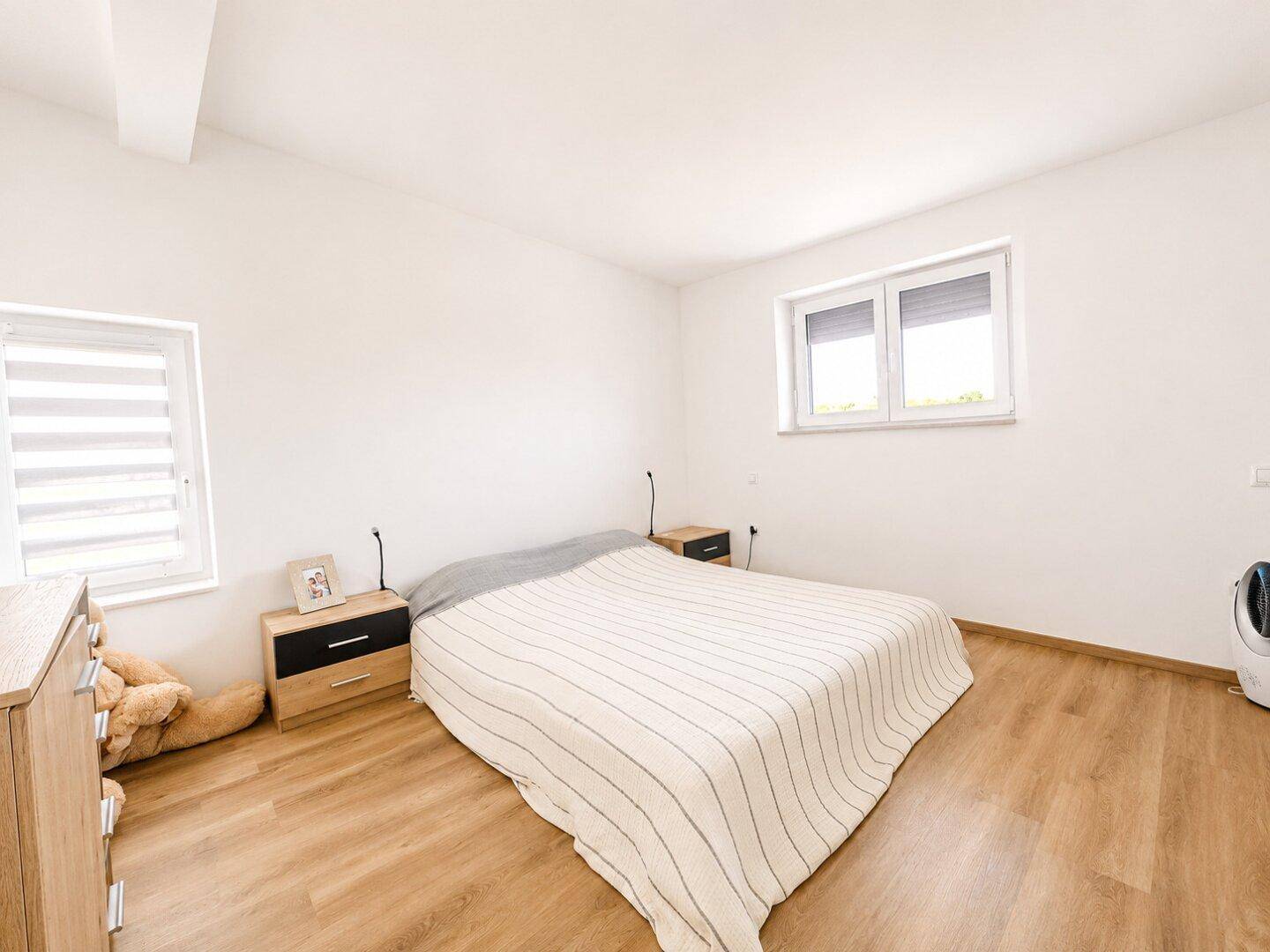 Helles Schlafzimmer mit Doppelbett, Nachttischen und zwei Fenstern für viel Tageslicht.