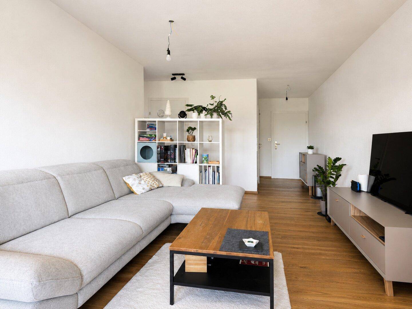 Helles Wohnzimmer mit gemütlicher Eckcouch, Holzboden und modernem Regal als Raumteiler.