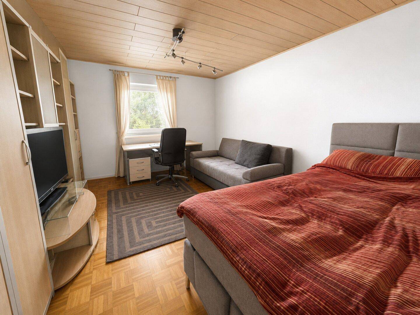 Schlafzimmer mit Bett, Schreibtisch und Sofa, ausgestattet mit Holzparkettboden.