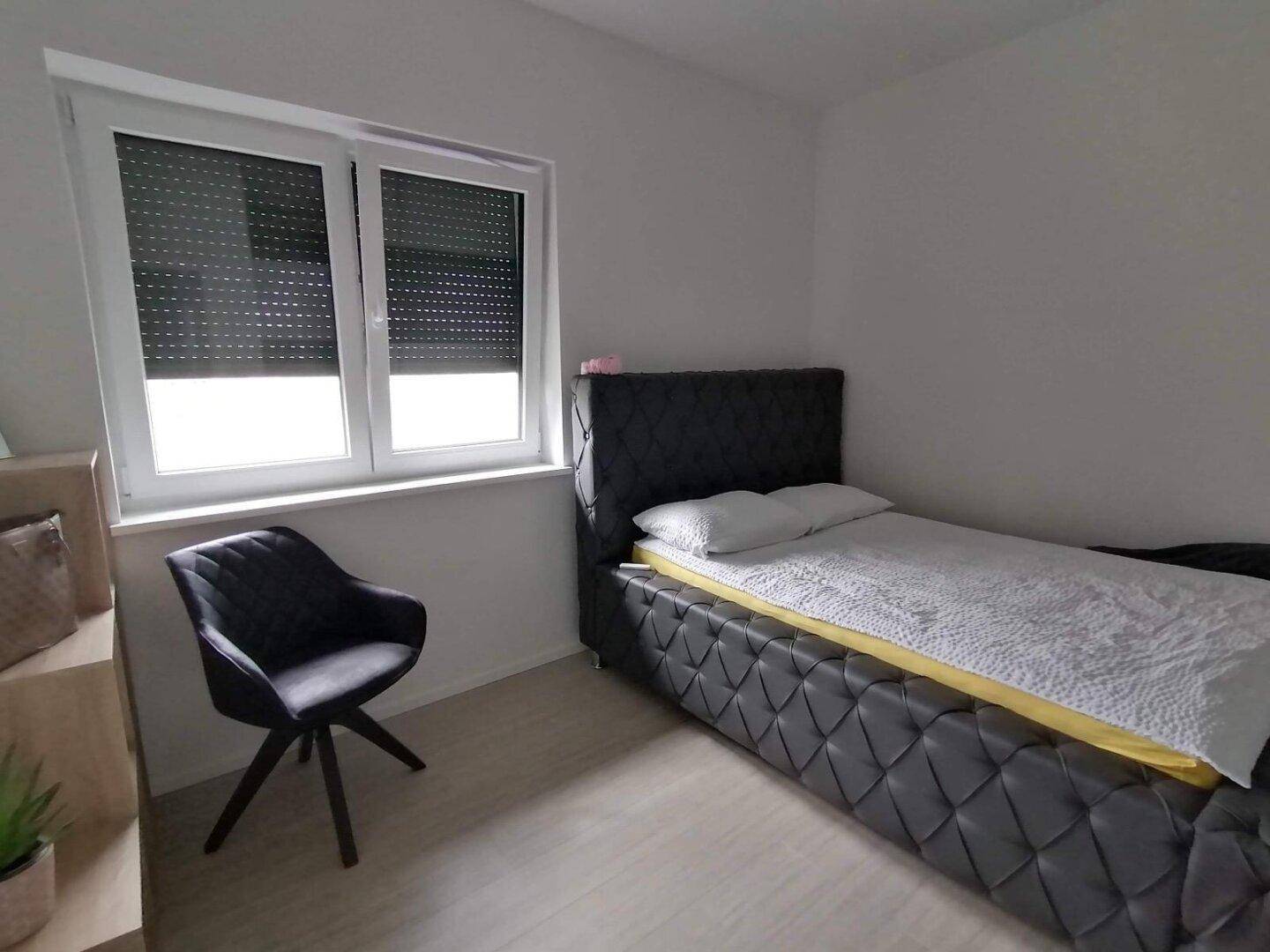 Helles Schlafzimmer mit Einzelbett, modernem Stuhl und Fenster mit Rollladen.