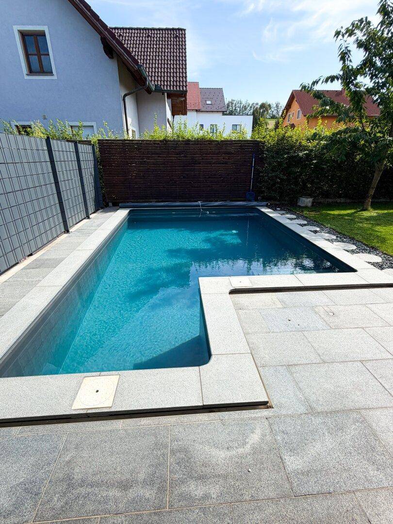 Moderner Außenpool mit grauen Pflastersteinen und umzäuntem Gartenbereich.