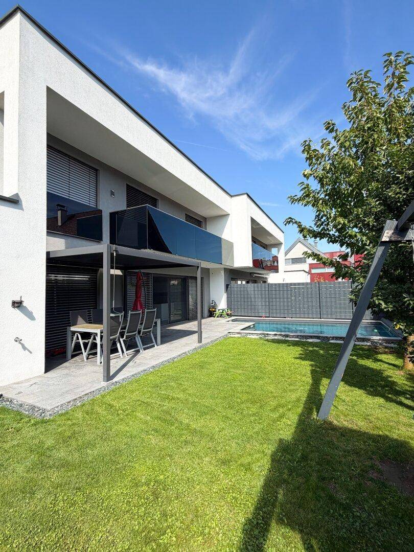 Moderne Hausfassade mit gepflegtem Rasen, überdachter Terrasse und Swimmingpool im Hintergrund.