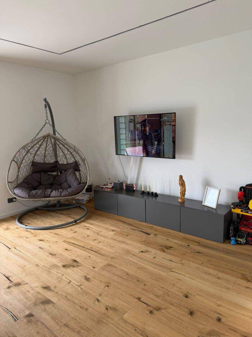 Modernes Wohnzimmer mit Holzdielenboden, Wandfernseher und einem gemütlichen Hängesessel.