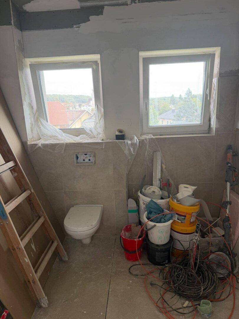Unfertiges Badezimmer mit gefliesten Wänden, Wand-WC und zwei Fenstern.