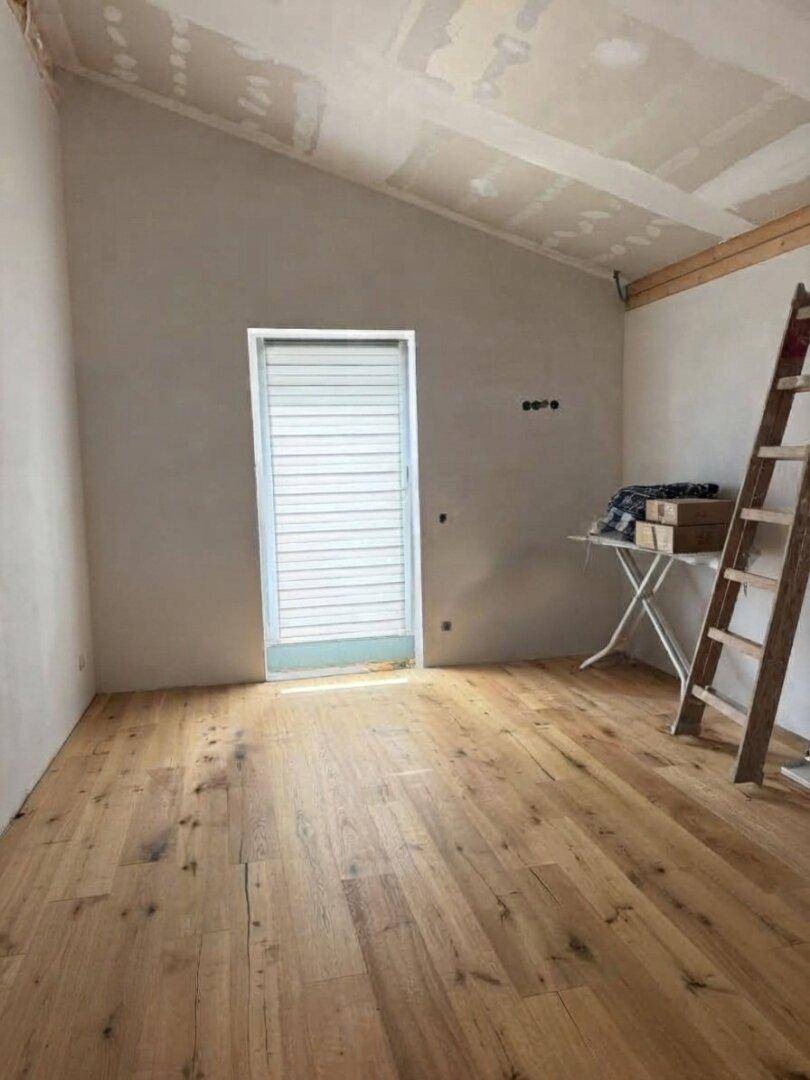 Unfertiger Raum mit Holzdielenboden, schräger Decke und Zugang nach draußen.