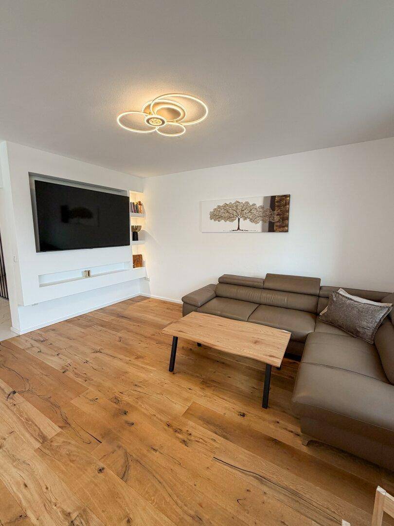 Gemütliches Wohnzimmer mit braunem Ecksofa, Holztisch und an der Wand montiertem Fernseher.