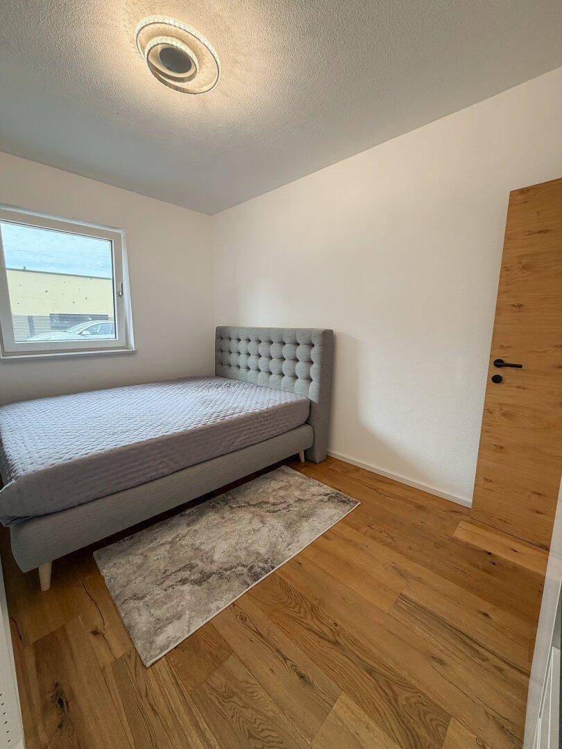 Schlafzimmer mit grauem Polsterbett, Holzboden und einem Fenster für natürliches Licht.