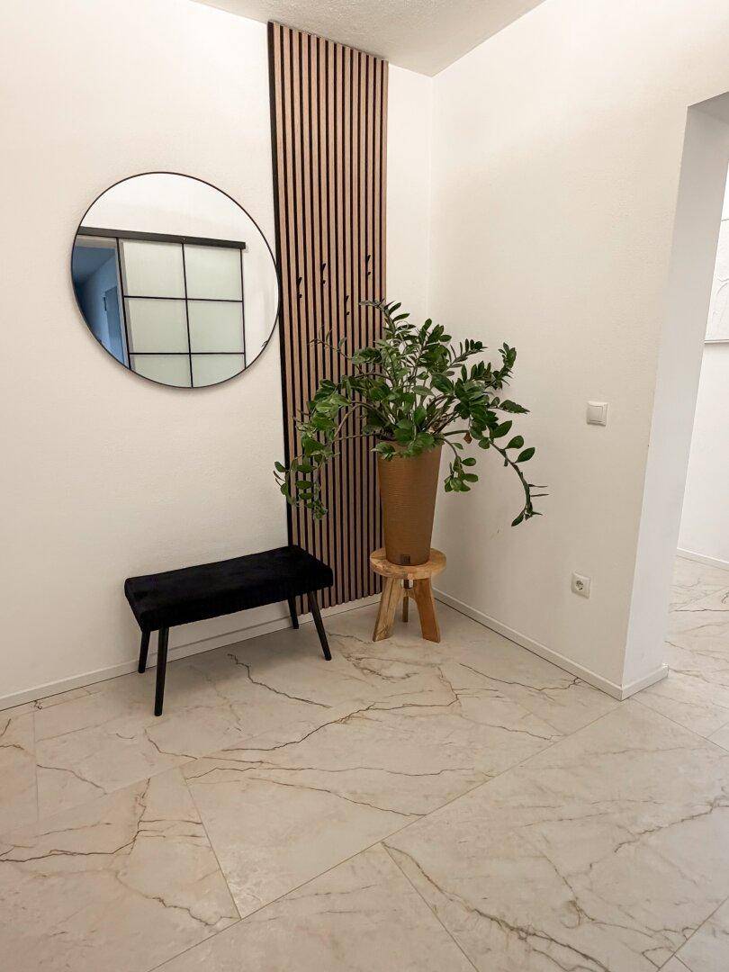 Heller Eingangsbereich mit Marmoroptik-Fliesenboden, rundem Spiegel und dekorativer Holzlamellenwand.