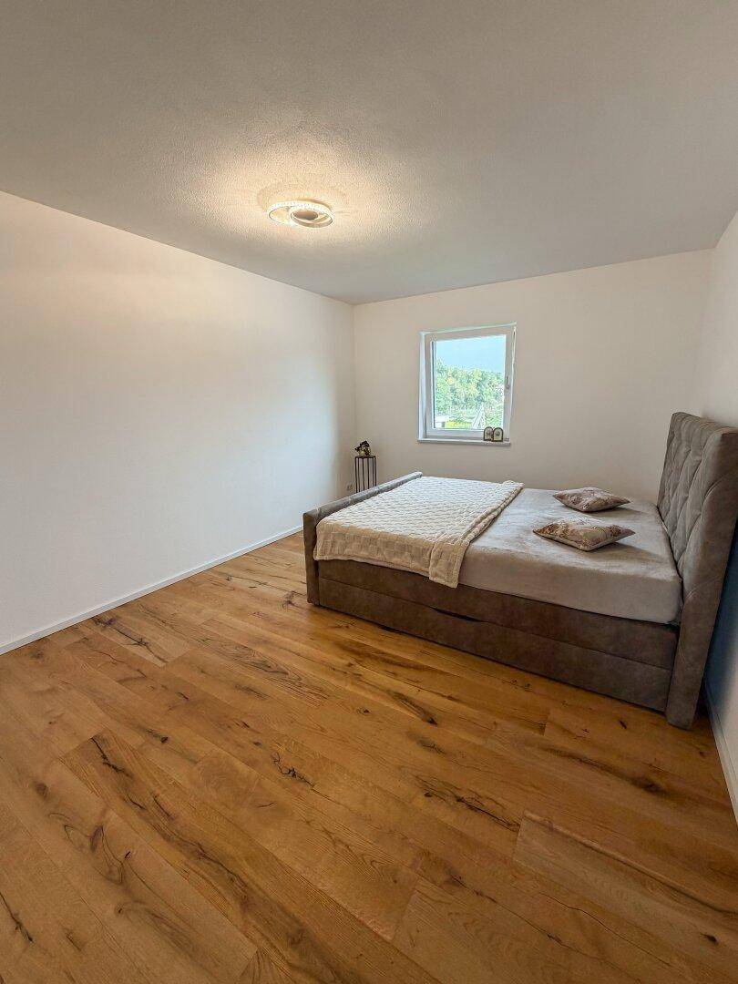 Schlafzimmer mit grauem Polsterbett, Holzboden und einem Fenster für natürliches Licht.