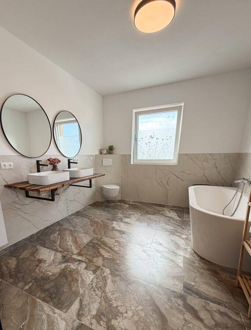 Großes Badezimmer mit freistehender Badewanne, Doppelwaschbecken und Marmoroptik-Fliesenboden.
