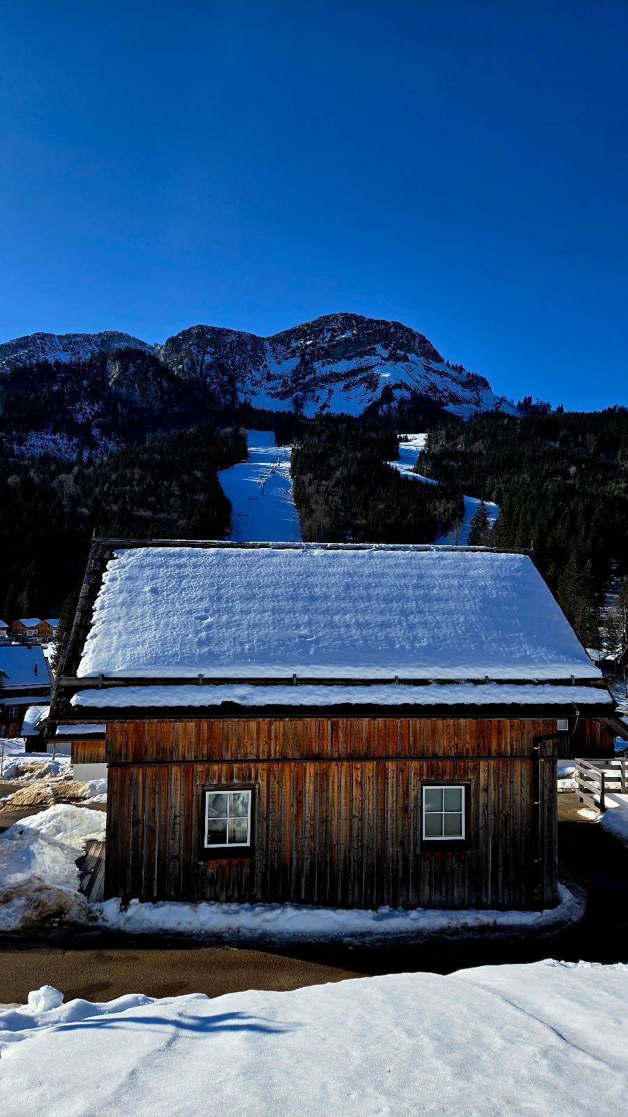 Traditionelles Holzhaus im Winter mit verschneitem Dach und Blick auf eine Skipiste.