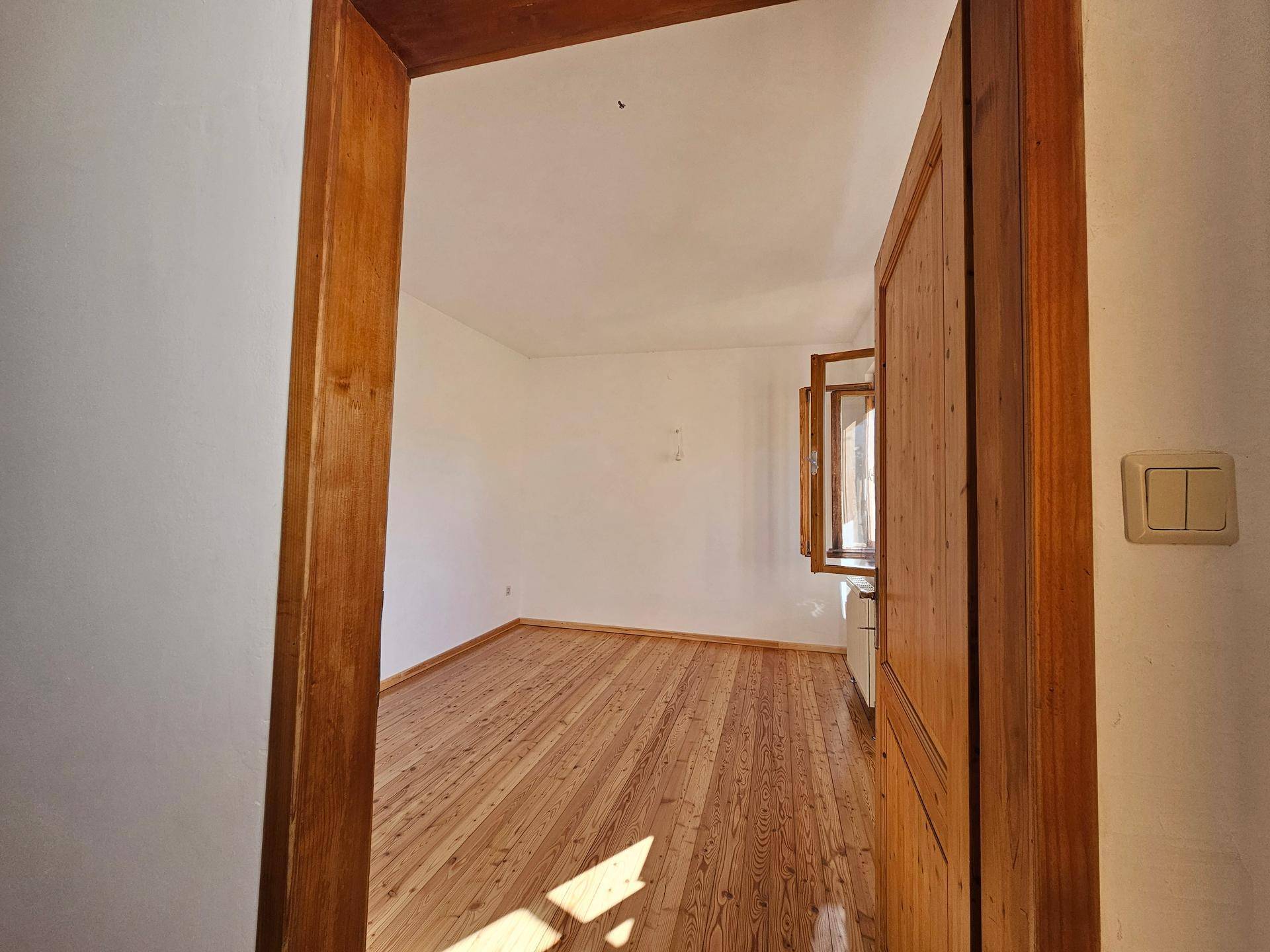 Helles Zimmer mit Holzdielenboden und weiß gestrichenen Wänden, ein Fenster ist sichtbar.