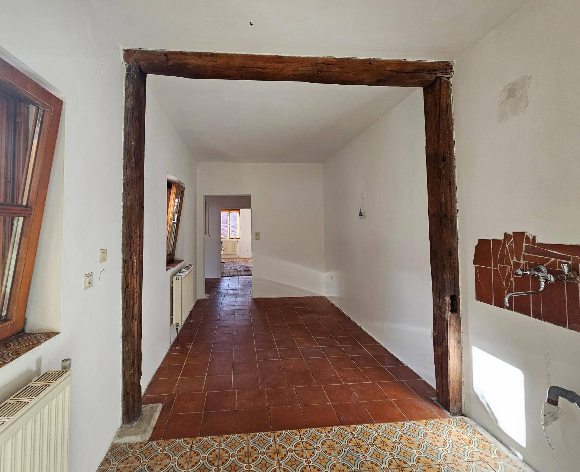 Innenraum mit sichtbaren Holzbalken und gemusterten Fliesen, führt zu weiteren Räumen im Haus.