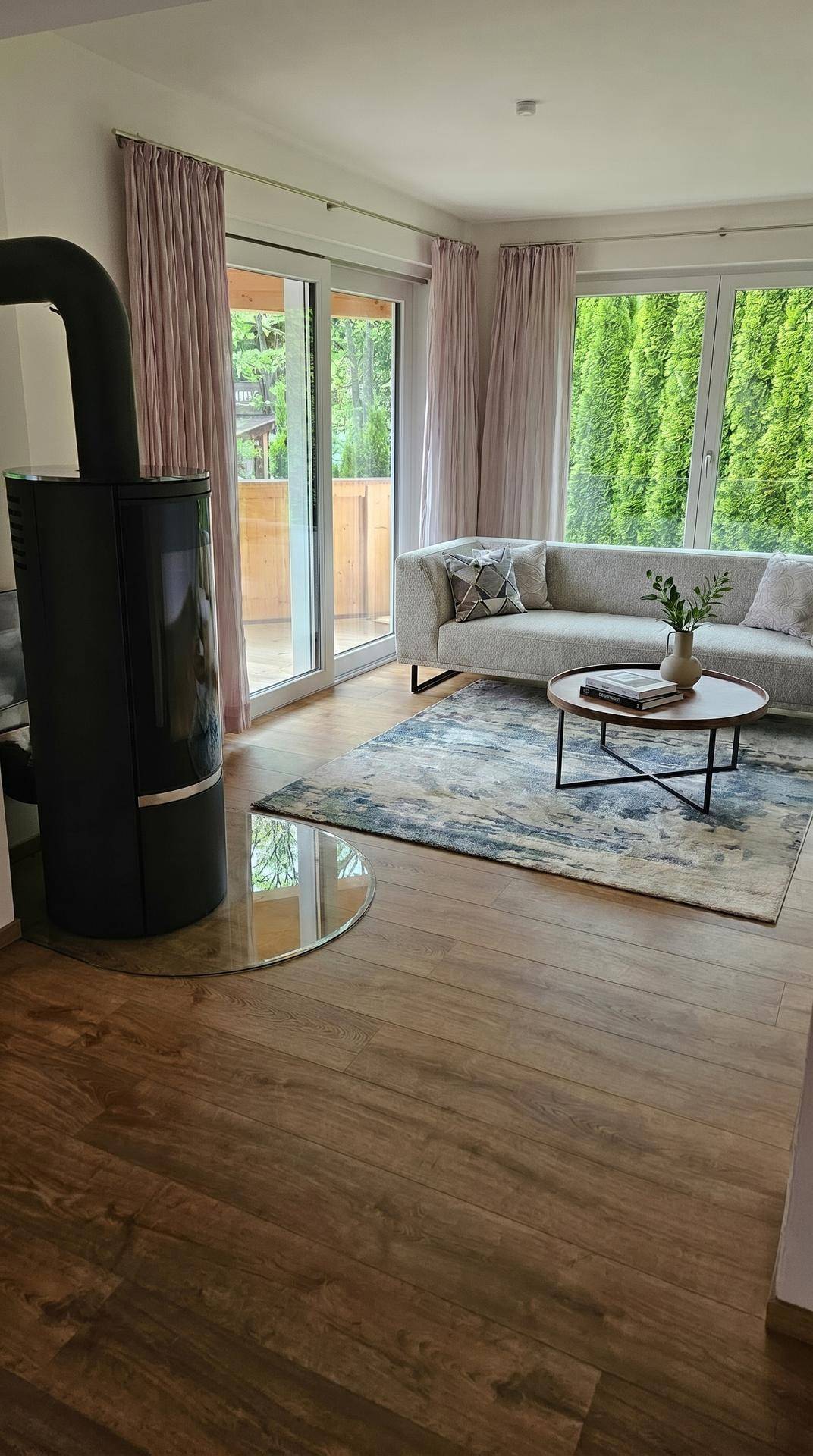 Modernes Wohnzimmer mit stilvollem Kaminofen, Holzboden und Blick ins Grüne durch große Fenster.
