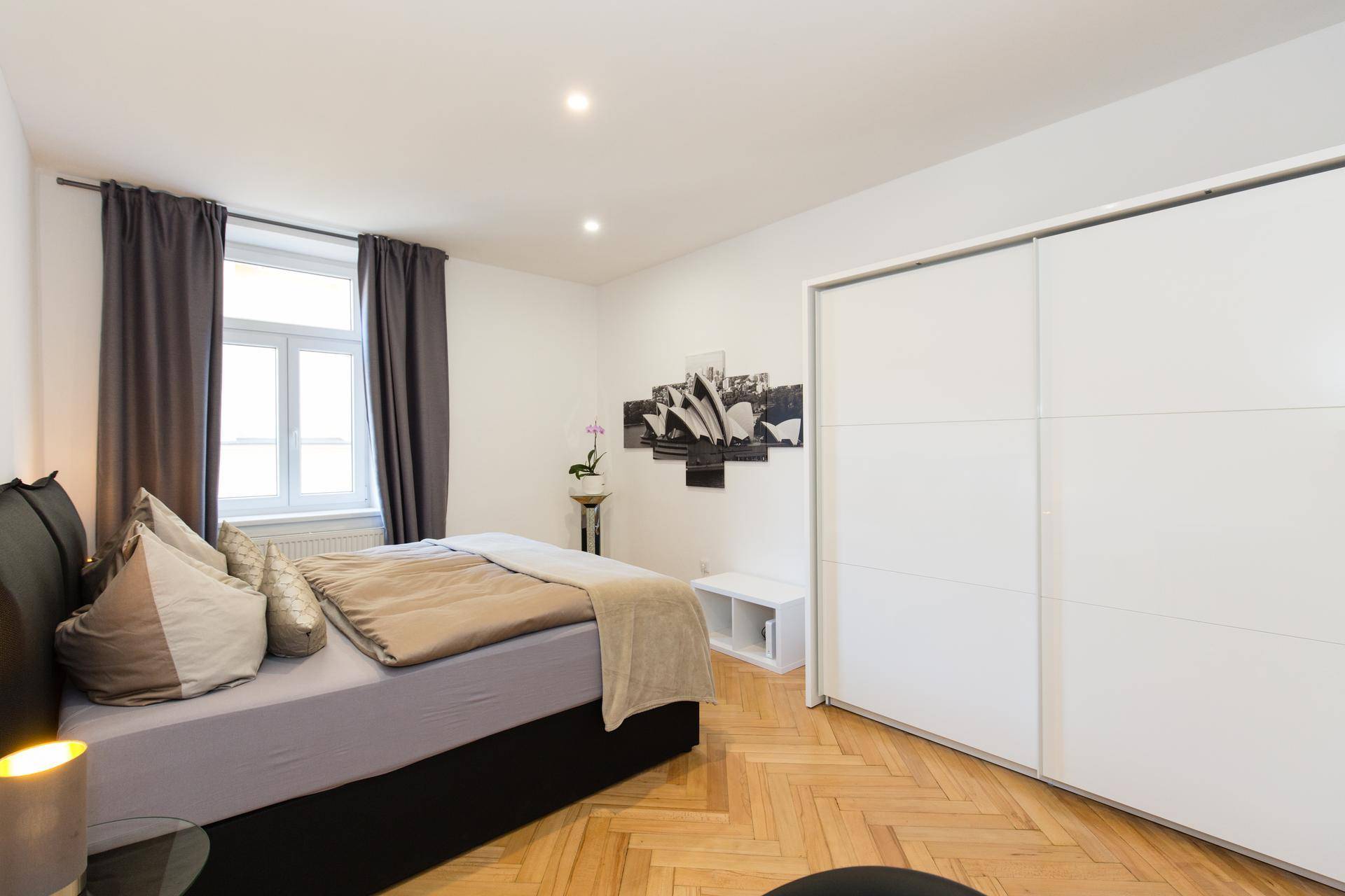 Helles Schlafzimmer mit Fischgrätparkett, großem Kleiderschrank und Fenster mit dunklen Vorhängen.