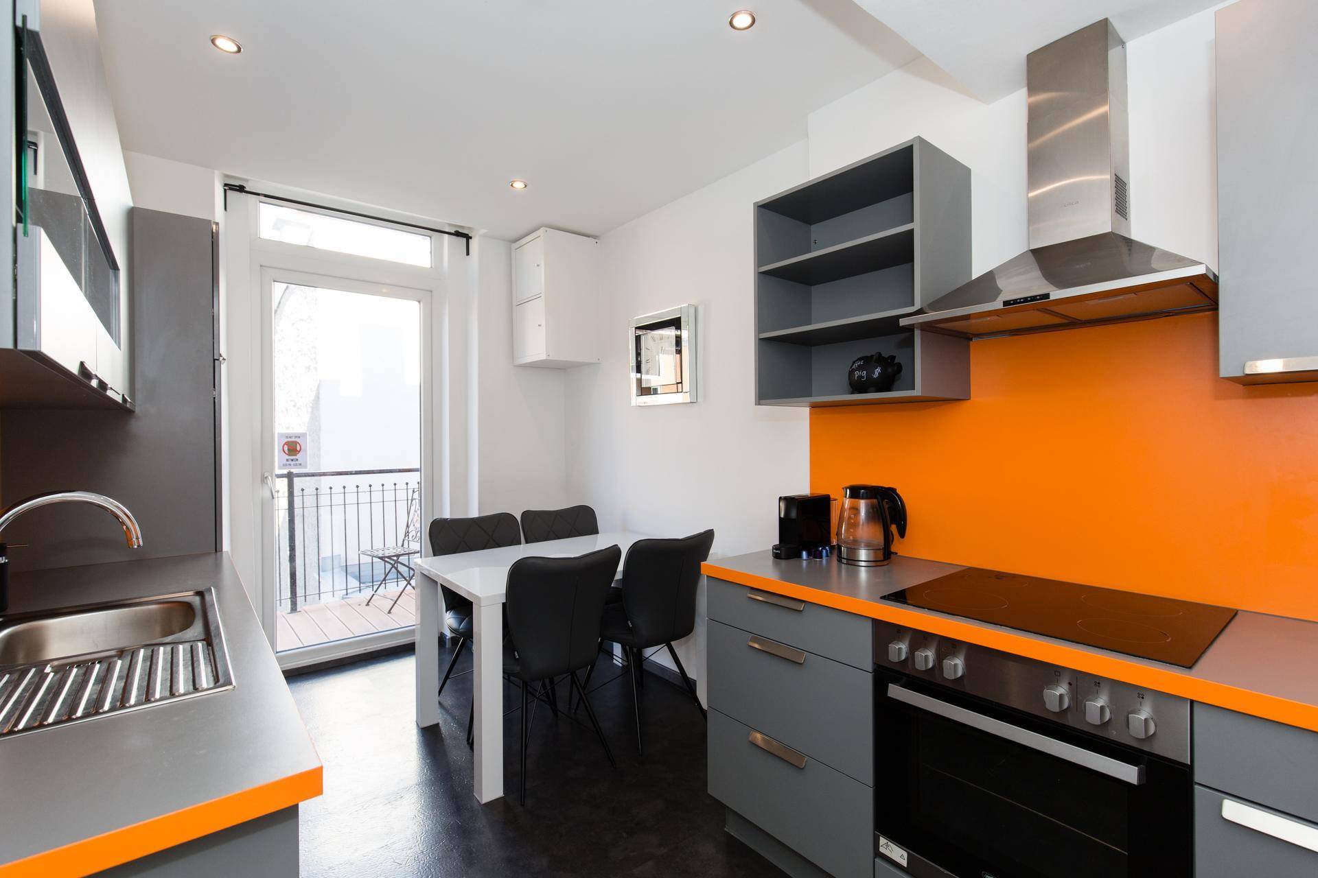 Moderne Küche mit grauen Schränken, orangefarbener Rückwand und Essbereich mit Balkonzugang.
