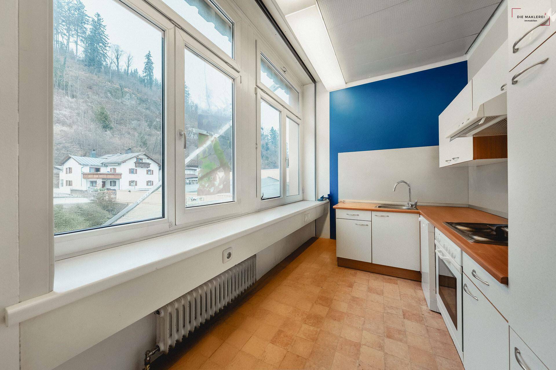 Kleine Küche mit weißen Schränken, blauer Wand und Blick auf die bewaldeten Hügel durch das Fenster.