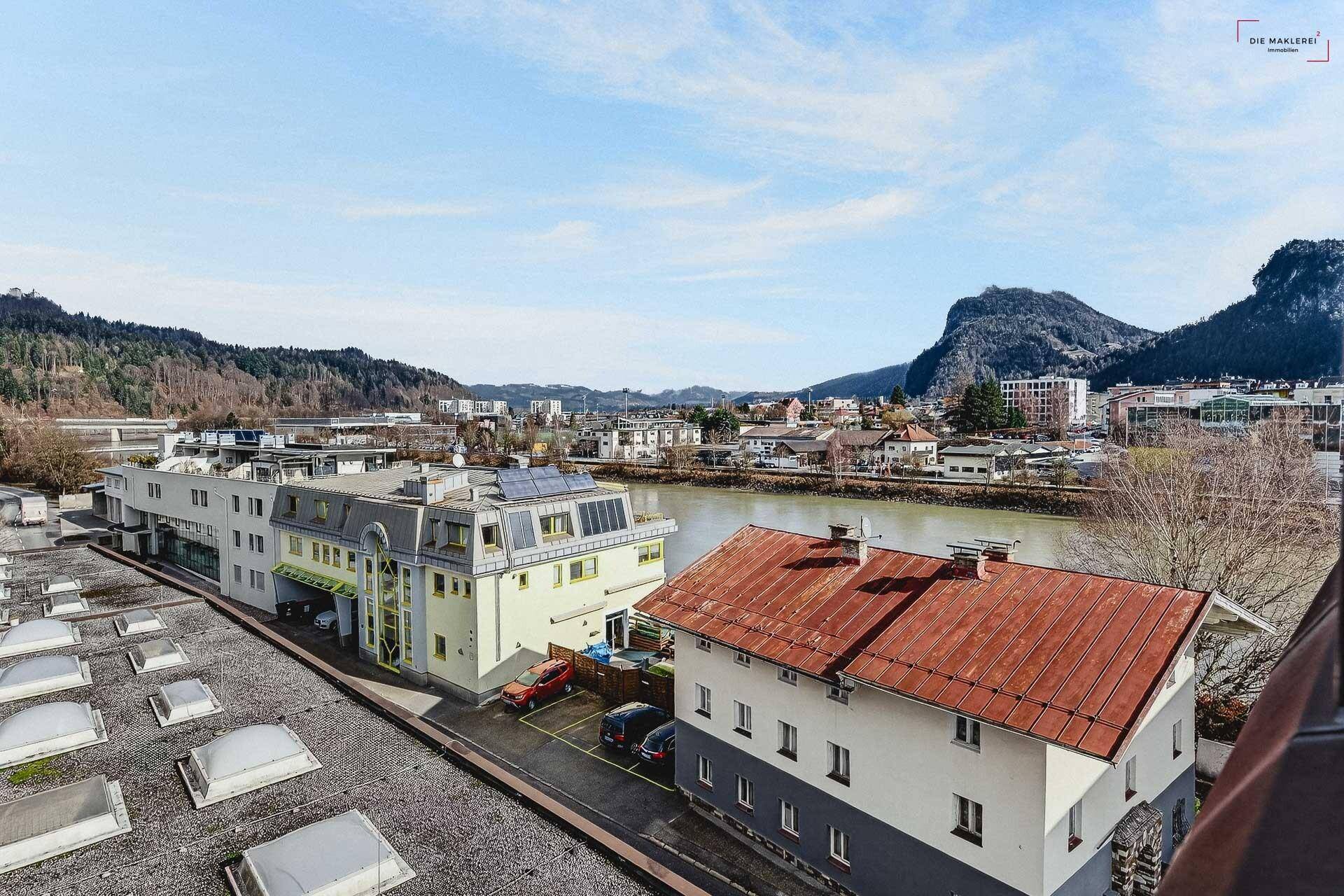Weitläufiger Blick über Gebäude, einen Fluss und die umliegende Berglandschaft unter klarem Himmel.