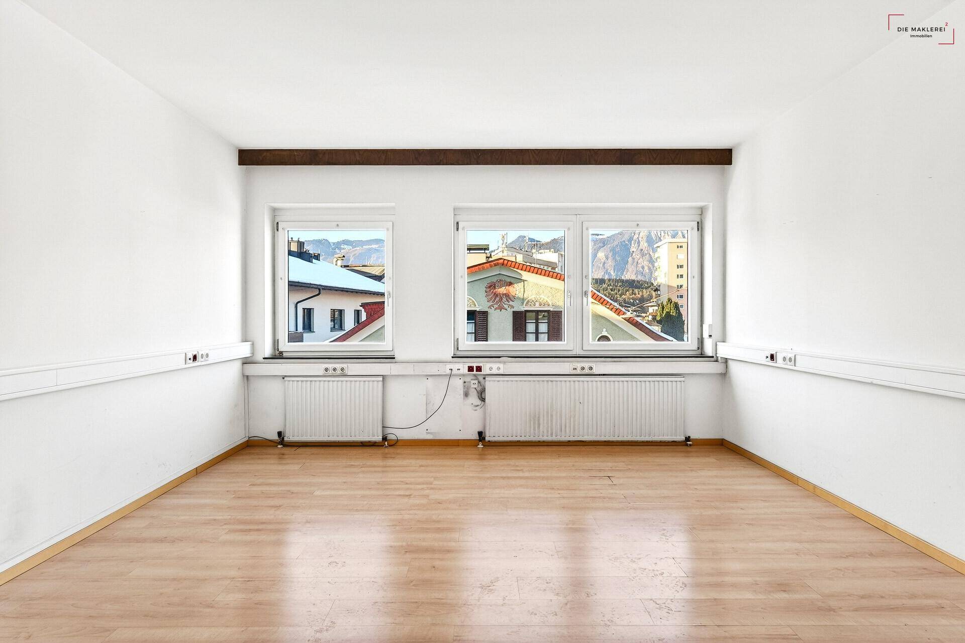 Heller Büroraum mit großen Fenstern, Laminatboden und Blick auf die umliegenden Gebäude und Berge.