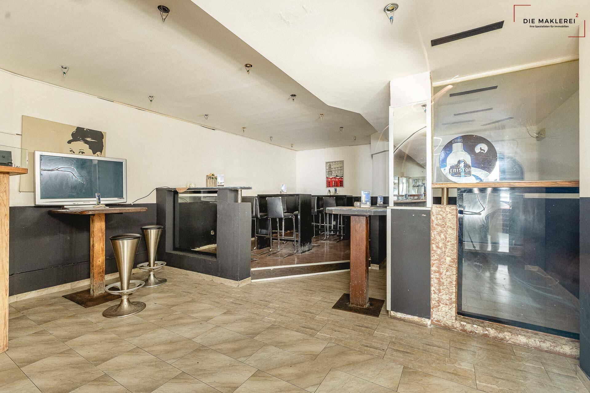 Sitzbereich einer Bar mit Tischen, Stühlen und einer erhöhten Plattform, alles auf einem gefliesten Boden.