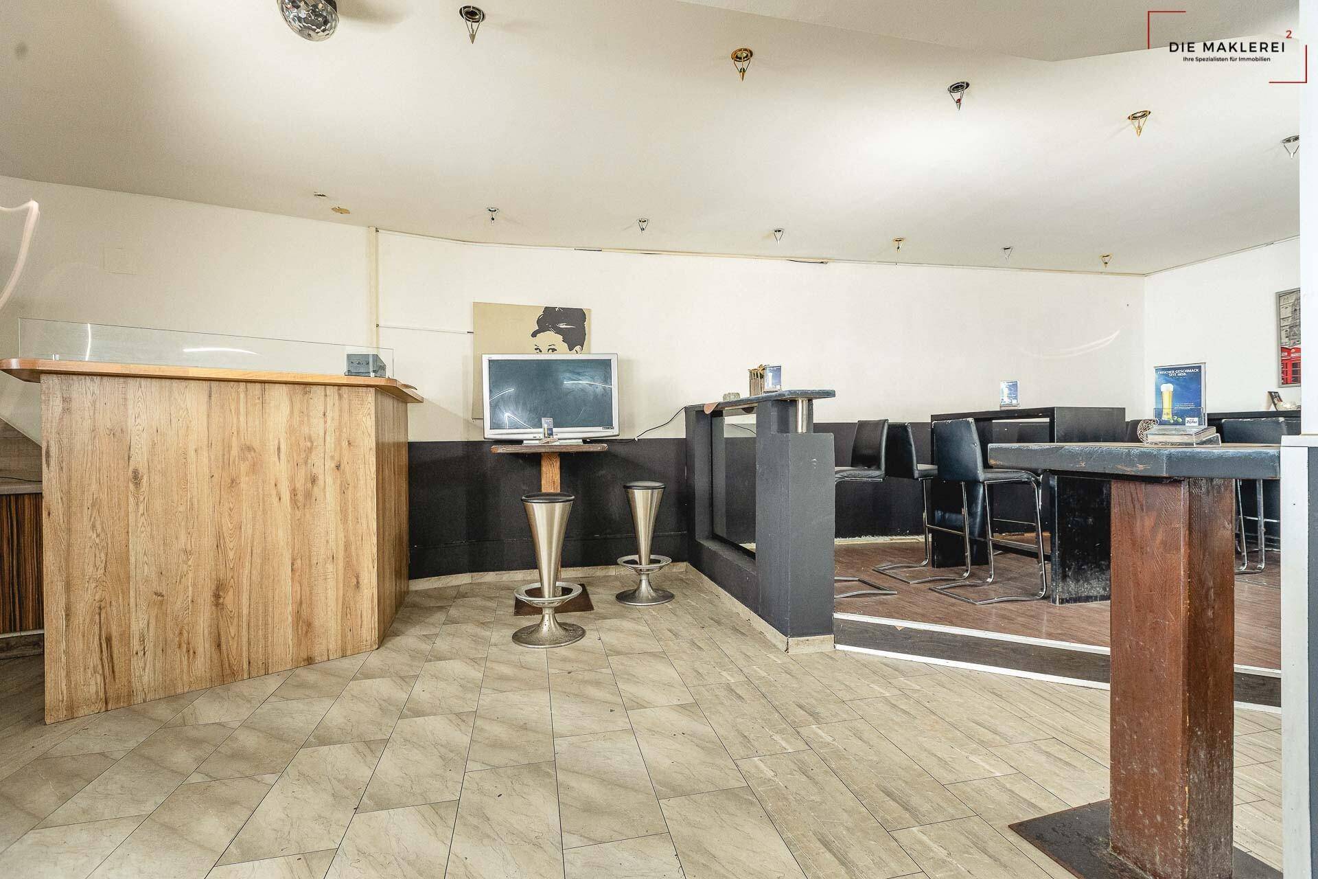 Sitzbereich einer Bar mit kleinerer Theke, Tischen und Stühlen, alles auf einem hell gefliesten Boden.