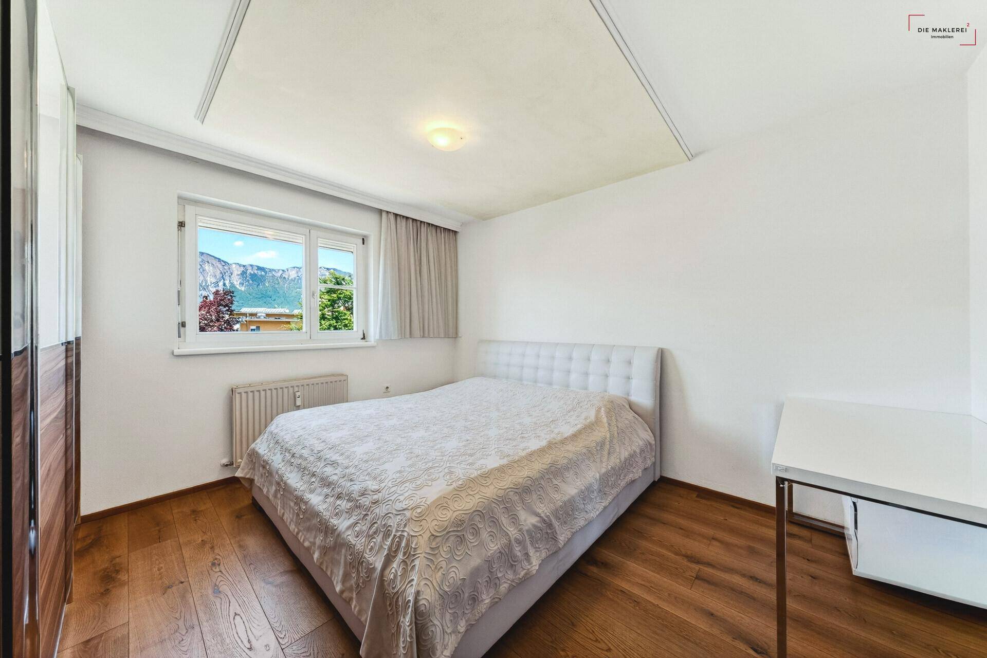 Helles Schlafzimmer mit weißem Bett, Holzboden und Bergblick aus dem Fenster.