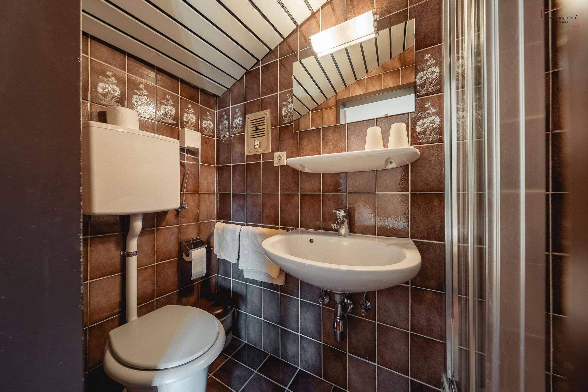 Kleines Badezimmer mit braunen Fliesen, Dusche und einem Waschbecken unter einem Spiegel.