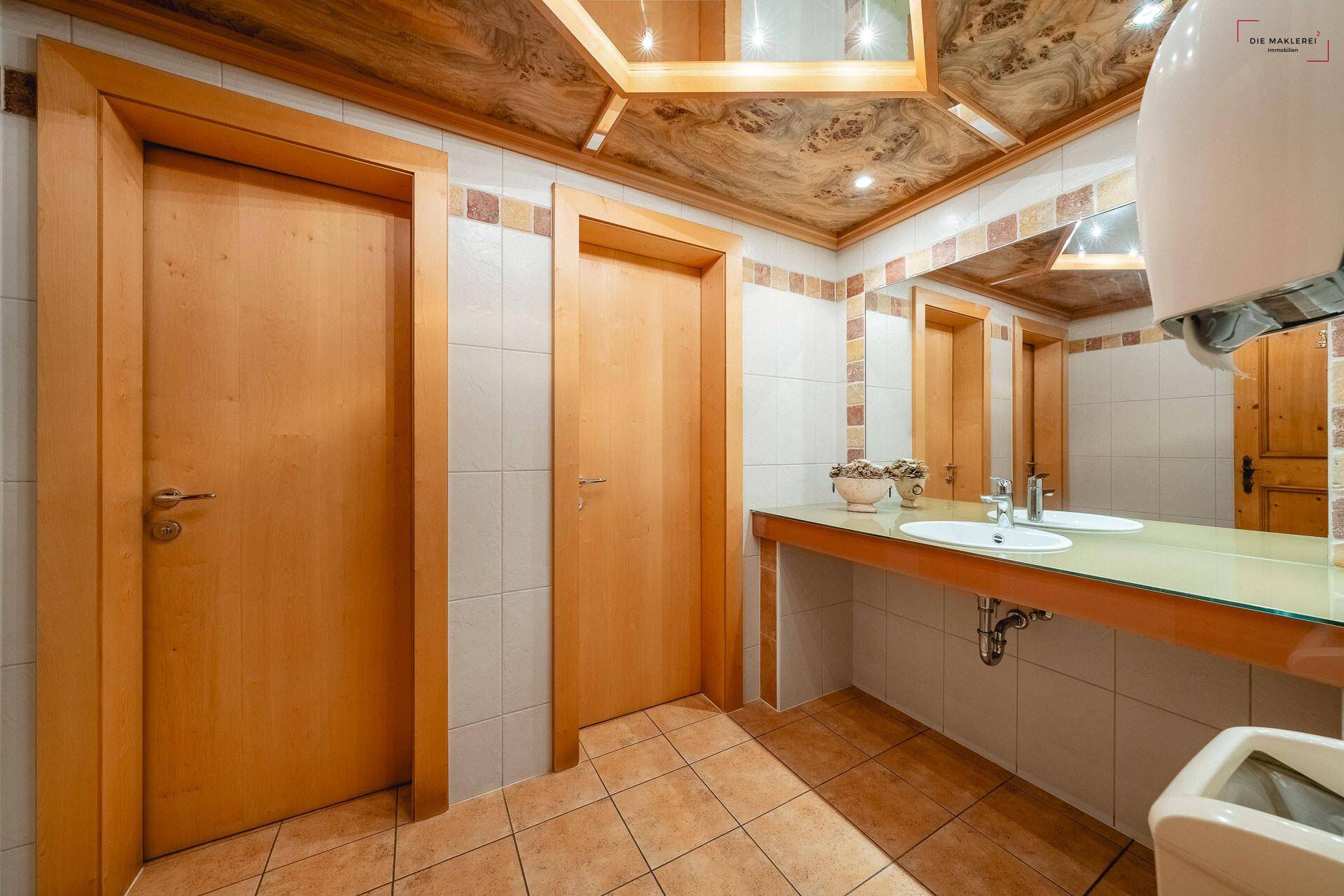 Helle Gästetoilette mit zwei Waschbecken, Holzdecke und neutralen Fliesen an den Wänden.
