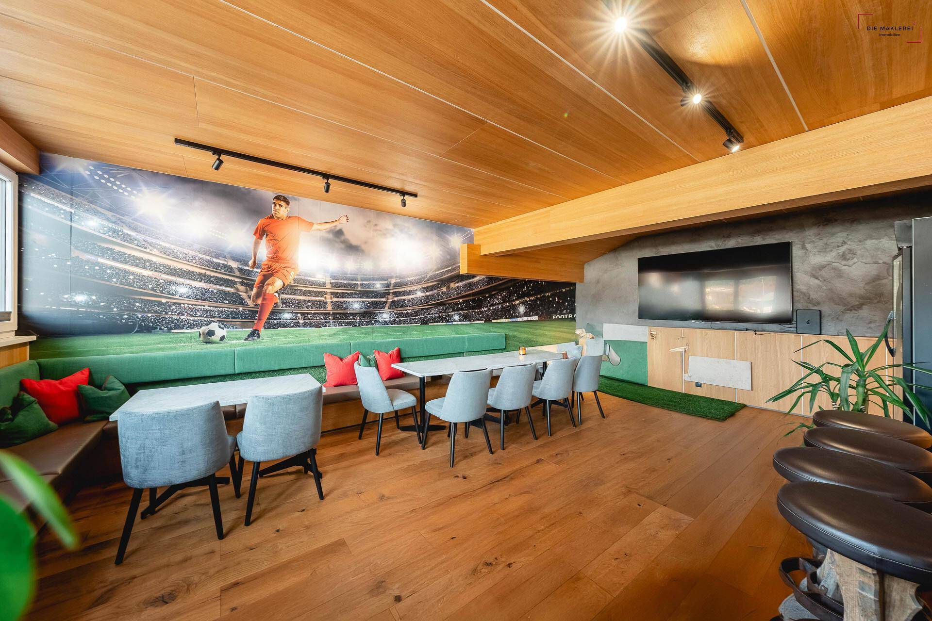 Moderner Gemeinschaftsraum mit Fußball-Wandbild, Holzboden und bequemen Sitzgelegenheiten.